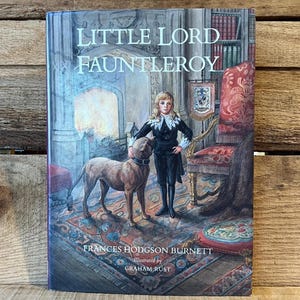 May include: A hardcover book titled "Little Lord Fauntleroy" with an illustration of a young person in a black suit and a dog. The book cover also features the author's name, Frances Hodgson Burnett, and the illustrator, Graham Rust.