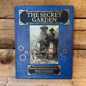 May include: A hardcover book titled "The Secret Garden" with a blue cover and gold accents. The cover features an illustration of a garden scene and the author's name, Frances Hodgson Burnett. The book's original illustrations are by Charles Robinson.