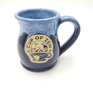 May include: A ceramic mug with a dark blue base and a lighter blue rim. The mug features a handle and a circular emblem with the words "Cup of Zen" and a cat meditating above a steaming cup of coffee.