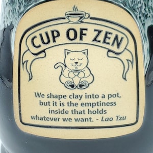 May include: A ceramic mug with a black and teal glaze. The mug features a tan label with the words "CUP OF ZEN" and a drawing of a cat holding a cup. The label also includes a quote by Lao Tzu.