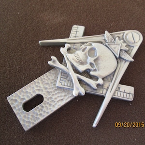 May include: A silver metal emblem featuring a skull, crossbones, a square, and a compass. The emblem has a textured surface and is likely a decorative item or a symbol of a fraternal organisation. The design incorporates Masonic symbols.
