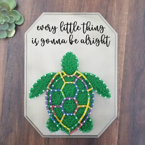May include: A string art piece featuring a green turtle design with colorful accents. The turtle is made of green string, with a geometric pattern in the center. The text "every little thing is gonna be alright" is above the turtle.