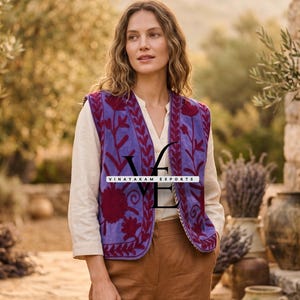May include: A woman wearing a purple embroidered vest with a white shirt and brown trousers. The vest features a V-neck and is adorned with red floral patterns. The text "VINAYAKAM EXPORTS" is displayed on the vest.
