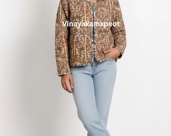 May include: A quilted jacket featuring a tan base with a repeating floral pattern in pink and green, long sleeves, and a round neckline. The jacket displays the text Vinayakamxpeot on the front. It is paired with light blue denim jeans and tan sandals.