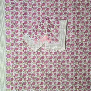 Op de afbeelding: Een witte stof met een herhalend patroon van kleine roze rozen met groene bladeren en stengels. Het textiel bevat een rand met een groter bloemmotief en een lichtgroen chevronpatroon. De tekst Pink City ArtCraft is zichtbaar.