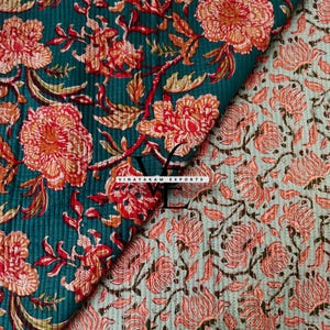 May include: A folded textile featuring two distinct patterns with a quilted texture. One side displays large orange and red floral motifs on a dark teal background, while the other shows smaller orange floral prints on a light green base. Text reads VINAYAKAM EXPORTS.