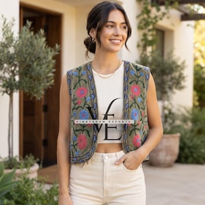 May include: An olive green sleeveless waistcoat featuring vibrant floral embroidery in shades of pink and blue along the front panels. The waistcoat is worn over a cream-coloured ribbed tank top and light-coloured trousers. The text VINAYAKAM EXPORTS is visible.