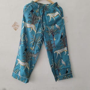 May include: Teal blue lounge trousers featuring a repeating pattern of leopards, toucans, and palm trees. The trousers have an elasticated waistband with a drawstring tie and white piping along the side seams and the bottom hem of the trouser legs.