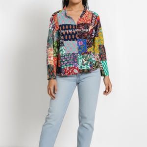 May include: A long-sleeved patchwork shirt with a mandarin collar. The shirt features a variety of colourful fabric patterns, including floral and geometric designs. The model is wearing light blue jeans and tan sandals. The text "inayakanmxpeg" is visible on the shirt.