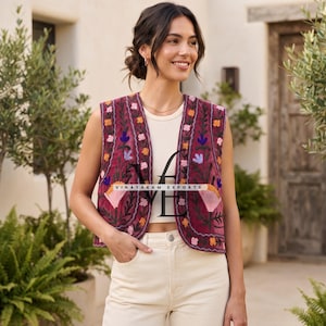 May include: A sleeveless maroon waistcoat featuring intricate floral embroidery in shades of orange, blue, and pink along the front panels. The garment is worn over a cream-coloured ribbed tank top and light-coloured jeans. The text VINAYAKAM EXPORTS is visible.
