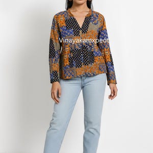 Puede incluir: Una blusa de manga larga con un diseño de patchwork en tonos naranja, azul y negro. La blusa tiene cuello en V y se combina con vaqueros azul claro. El texto "Vinayakamxpeot" es visible en la blusa.