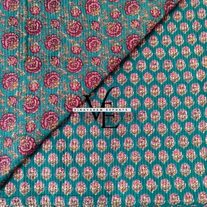 May include: A teal fabric featuring a quilted texture and a repeating floral pattern in shades of pink and white. The fabric is folded to show two different floral print scales. The text VINAYAKAM EXPORTS is printed in black in the center.