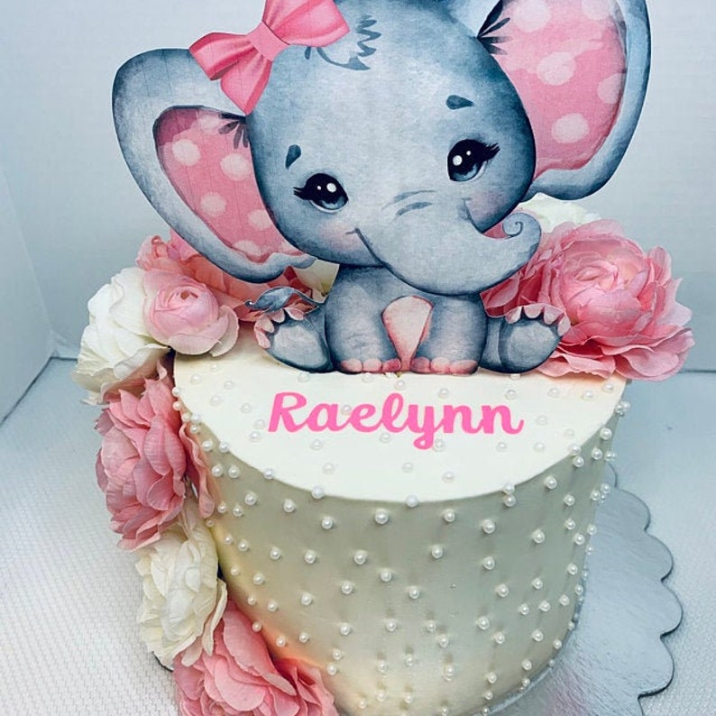 May include: A white cake decorated with white pearl sprinkles and pink flowers. A gray elephant with pink ears and a pink bow is on top of the cake. The name "Raelynn" is written in pink frosting on the cake.