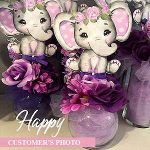 May include: Purple and pink floral centerpieces with a gray elephant figurine wearing a pink flower crown. The text "Happy" is visible in the image.
