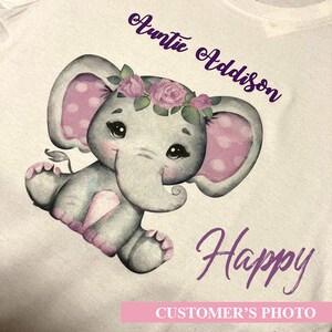 May include: A white t-shirt with a purple elephant illustration wearing a pink flower crown. The text "Auntie Addison" is written in purple above the elephant and "Happy" is written in purple below the elephant. The text "CUSTOMER'S PHOTO" is written in pink at the bottom of the image.