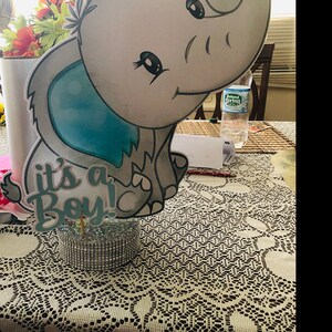 Puede incluir: Una figurita de elefante gris y azul con una pajarita azul y el texto "it's a Boy!" sobre una base plateada brillante.
