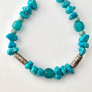 May include: A turquoise and silver beaded bracelet with a wire closure. The bracelet features turquoise stones of various shapes and sizes, as well as two silver beads with geometric designs.