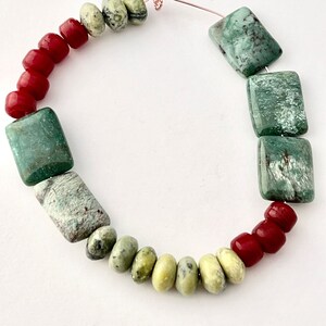May include: A handmade necklace featuring a variety of beads. The necklace includes rectangular turquoise-colored beads, red beads, and oval-shaped green beads. The beads are strung together on a copper wire, creating a unique and colorful accessory.