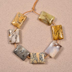 May include: A strand of seven rectangular, multi-colored agate beads with brown, white, and yellow patterns. The beads are strung on a brown cord.