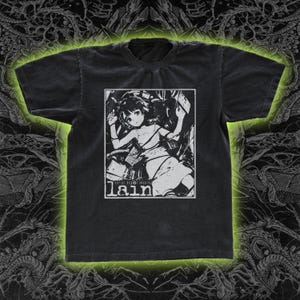 May include: Dark gray t-shirt featuring a black and white graphic of an anime character. The graphic includes the word "lain" and the text "serial experiments lain". The shirt has a crew neck and short sleeves.