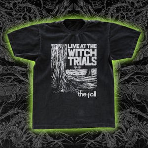May include: Black t-shirt featuring a white graphic with the text "LIVE AT THE WITCH TRIALS" above an illustration of a tree and the text "the fall" below. The shirt has a crew neck and short sleeves.
