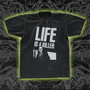 May include: Dark gray t-shirt with the text "LIFE IS A KILLER" in white block letters. Below the text is a white graphic of a man in a suit holding a cigarette and a white outline of a state. The shirt has a vintage, worn look.