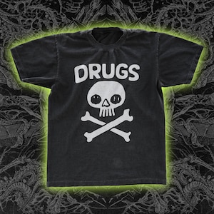 May include: Black t-shirt with the word "DRUGS" in white block letters above a white skull and crossbones graphic. The shirt has a crew neck and short sleeves. The t-shirt has a vintage, worn look.