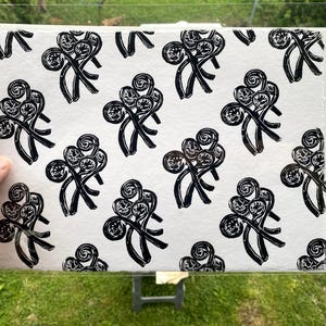 May include: A black and white print featuring a repeating pattern of stylized fern fronds. The print is held up in front of a green grassy background. The fern fronds are black and the background is white.