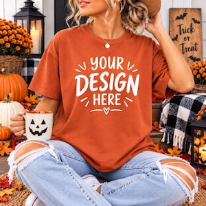 Könnte beinhalten: Ein rostfarbenes Kurzarm-T-Shirt mit dem weißen Schriftzug "YOUR DESIGN HERE" und einem kleinen Herz-Symbol, kombiniert mit einer hellblauen, zerrissenen Jeans und einem weißen Becher mit schwarzem Kürbisgesicht.