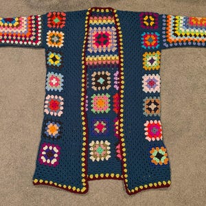 May include: A long-sleeved open-front cardigan made of dark blue yarn featuring a patchwork design of multi-colored granny squares. The garment has a deep blue base color with vibrant square motifs in shades of pink, yellow, orange, and green.