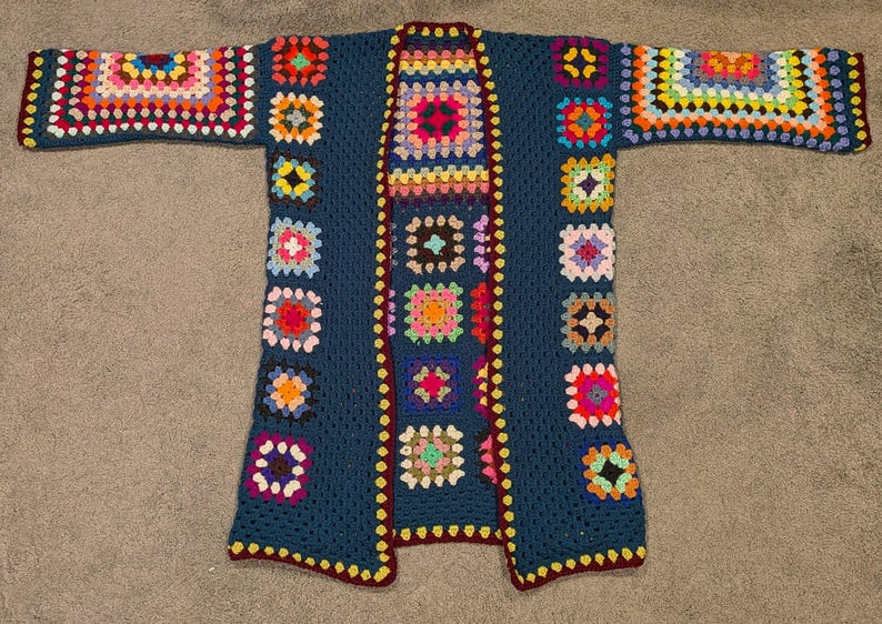 May include: A long-sleeved open-front cardigan made of dark blue yarn featuring a patchwork design of multi-colored granny squares. The garment has a deep blue base color with vibrant square motifs in shades of pink, yellow, orange, and green.