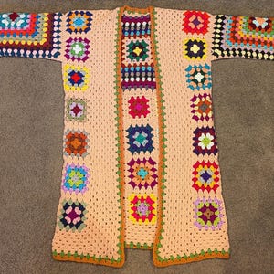 May include: A long-sleeved cardigan made of crocheted granny squares in a variety of vibrant colors including red, blue, yellow, green, and purple, set against a light peach-colored background with an orange border along the front opening.