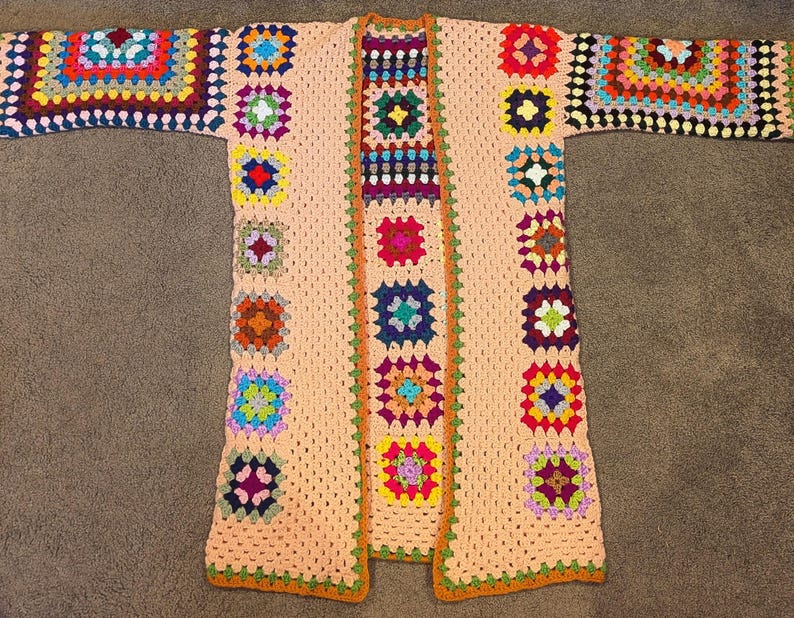 May include: A long-sleeved cardigan made of crocheted granny squares in a variety of vibrant colors including red, blue, yellow, green, and purple, set against a light peach-colored background with an orange border along the front opening.