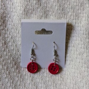 May include: A pair of handmade earrings featuring bright red button charms. The button earrings have silver-colored fish hook ear wires. The earrings are displayed on a white earring card.