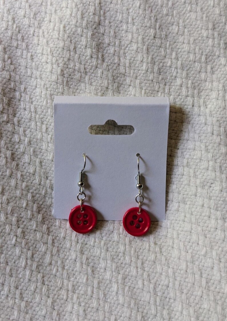 May include: A pair of handmade earrings featuring bright red button charms. The button earrings have silver-colored fish hook ear wires. The earrings are displayed on a white earring card.