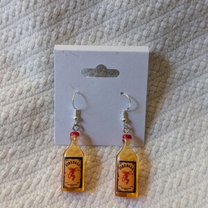 May include: A pair of novelty earrings featuring miniature Fireball whiskey bottle charms. The charms are amber-colored with red bottle caps and silver-tone ear wires. Each bottle has a label with a red dragon logo.