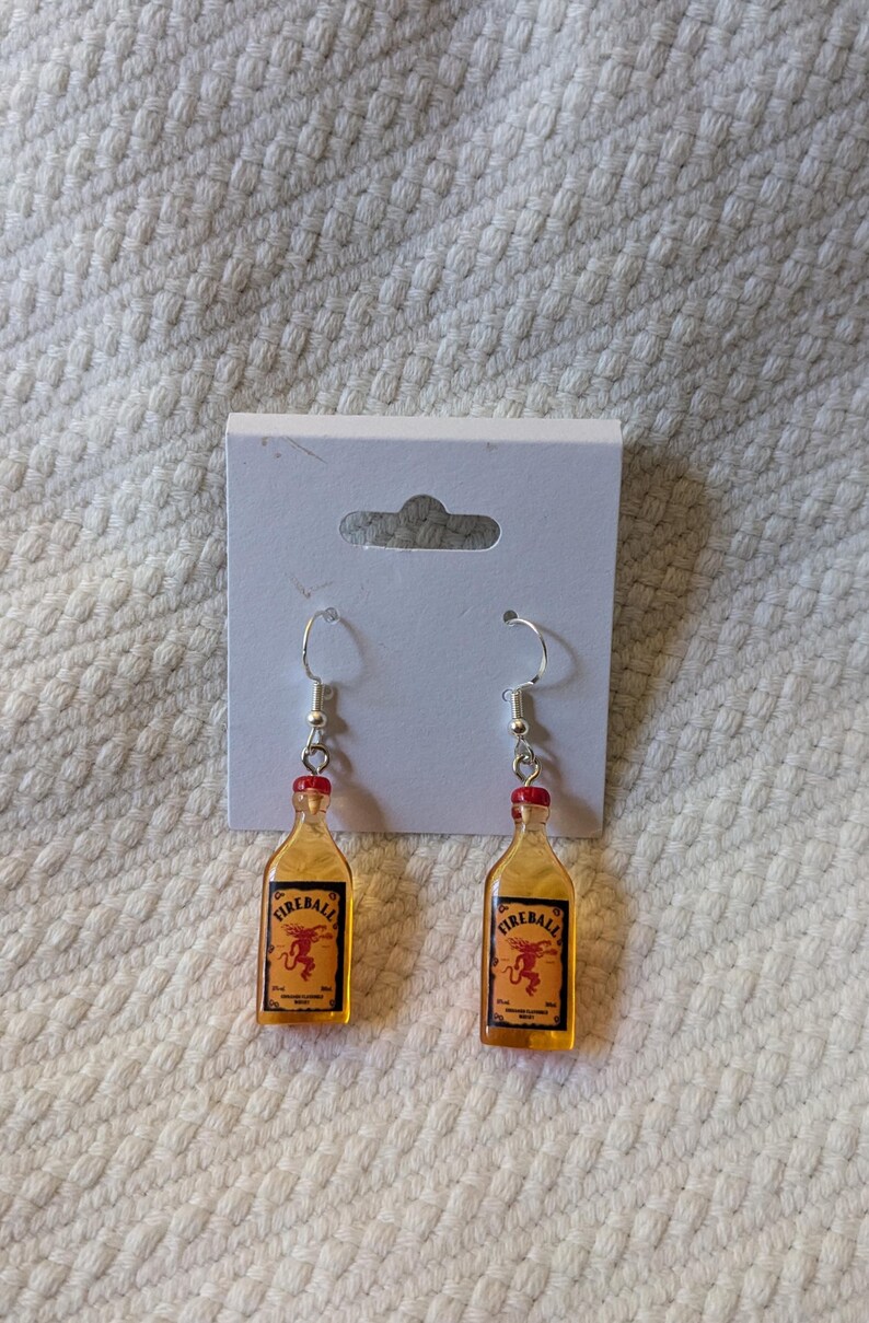 May include: A pair of novelty earrings featuring miniature Fireball whiskey bottle charms. The charms are amber-colored with red bottle caps and silver-tone ear wires. Each bottle has a label with a red dragon logo.