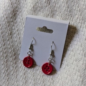 May include: A pair of handmade earrings featuring bright red button charms. The buttons have four holes and are attached to silver-colored fish hook ear wires. The earrings are displayed on a white earring card against a textured white background.