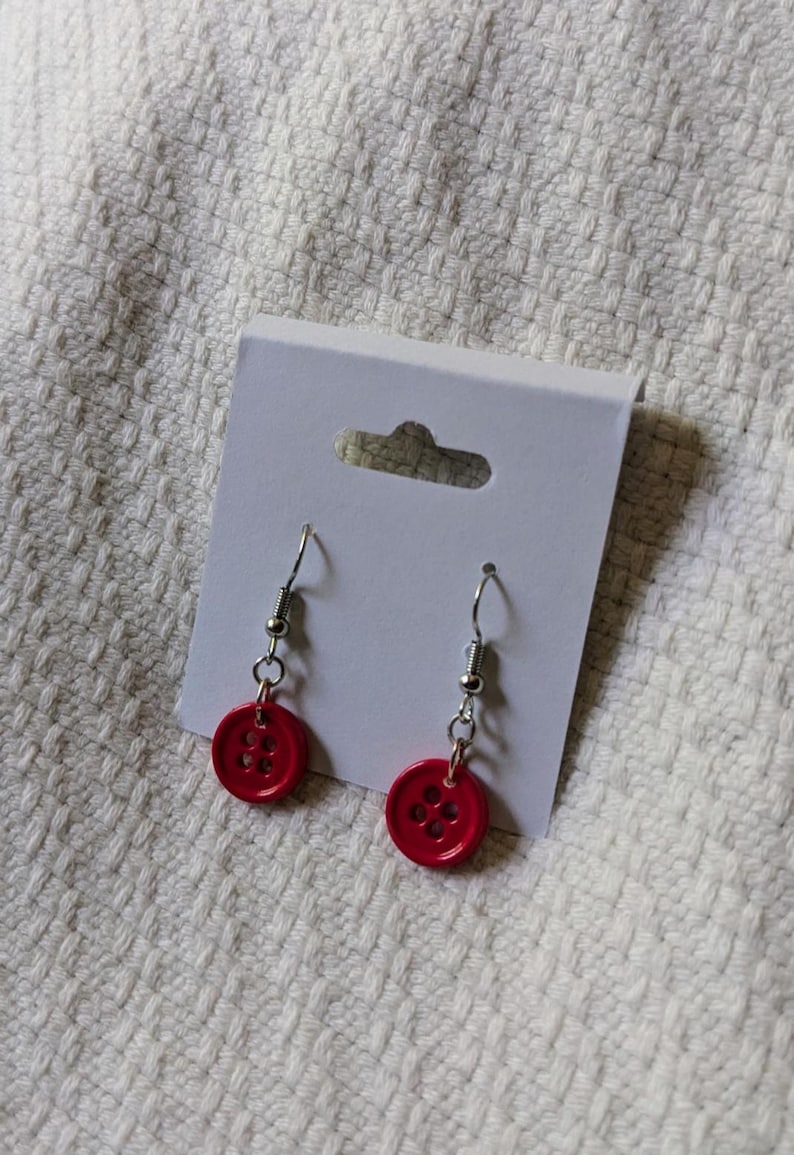 May include: A pair of handmade earrings featuring bright red button charms. The buttons have four holes and are attached to silver-colored fish hook ear wires. The earrings are displayed on a white earring card against a textured white background.