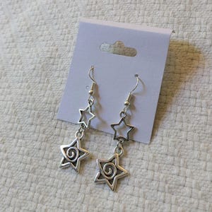 Puede incluir: Pendientes de estrella plateados con un diseño en espiral en la estrella inferior. Los pendientes cuelgan de un gancho plateado y se exhiben en una tarjeta blanca. Los pendientes miden aproximadamente 5 cm de largo.