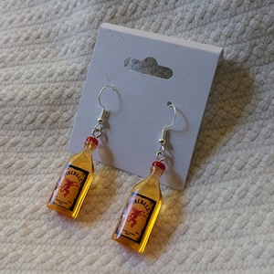 May include: A pair of dangle earrings featuring miniature Fireball whiskey bottle charms. The charms are amber-colored with red bottle caps and silver-tone ear wires. Each bottle has a label with the word "Fireball" and a red dragon graphic.