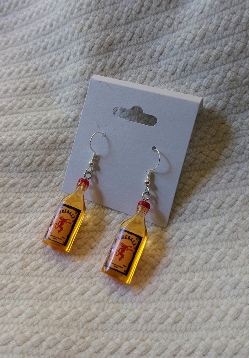 May include: A pair of dangle earrings featuring miniature Fireball whiskey bottle charms. The charms are amber-colored with red bottle caps and silver-tone ear wires. Each bottle has a label with the word "Fireball" and a red dragon graphic.