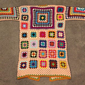 May include: A long-sleeved cardigan made from a patchwork of multicolored granny squares. The garment features a light peach-colored yarn border connecting the squares, with a large central square on the back and smaller squares forming the sleeves.