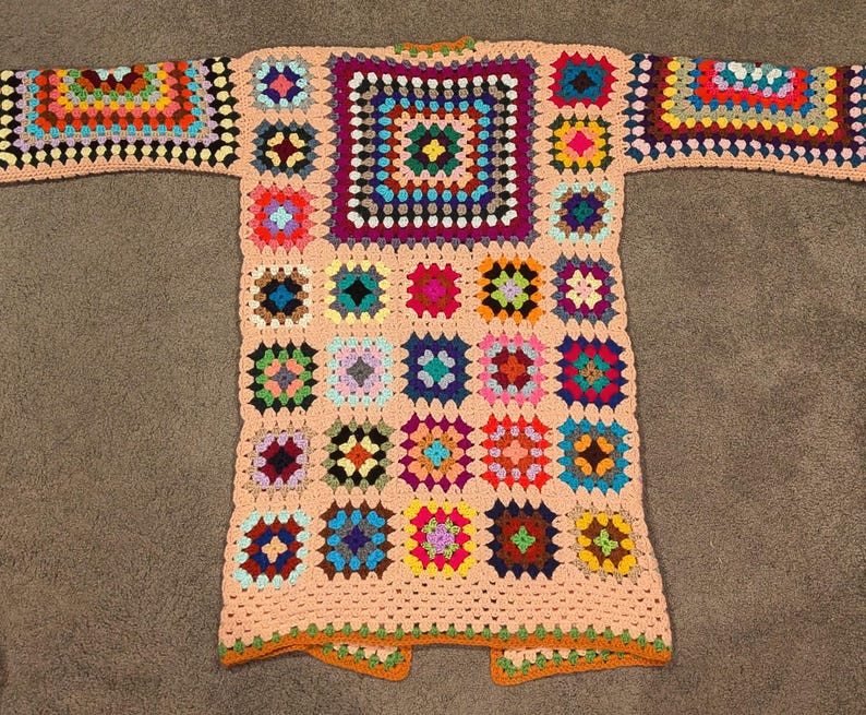 May include: A long-sleeved cardigan made from a patchwork of multicolored granny squares. The garment features a light peach-colored yarn border connecting the squares, with a large central square on the back and smaller squares forming the sleeves.