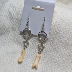May include: A pair of silver-toned drop earrings. Each earring has a small bone charm, a star-shaped charm, and a silver hoop. The earrings are presented on a white earring card.