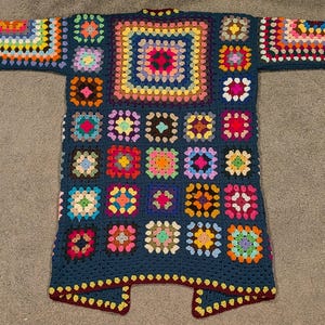 May include: A long-sleeved cardigan made from dark blue yarn featuring a patchwork of multi-colored granny squares. The garment displays a variety of vibrant hues including pink, yellow, orange, green, and purple arranged in a repeating square pattern.