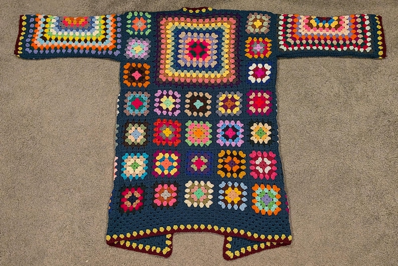 May include: A long-sleeved cardigan made from dark blue yarn featuring a patchwork of multi-colored granny squares. The garment displays a variety of vibrant hues including pink, yellow, orange, green, and purple arranged in a repeating square pattern.