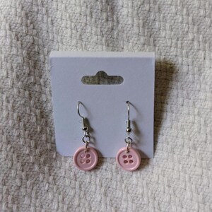 May include: A pair of button earrings. Each earring features a light pink button with four holes, suspended from a silver-colored fish hook. The earrings are displayed on a white card against a textured white background.