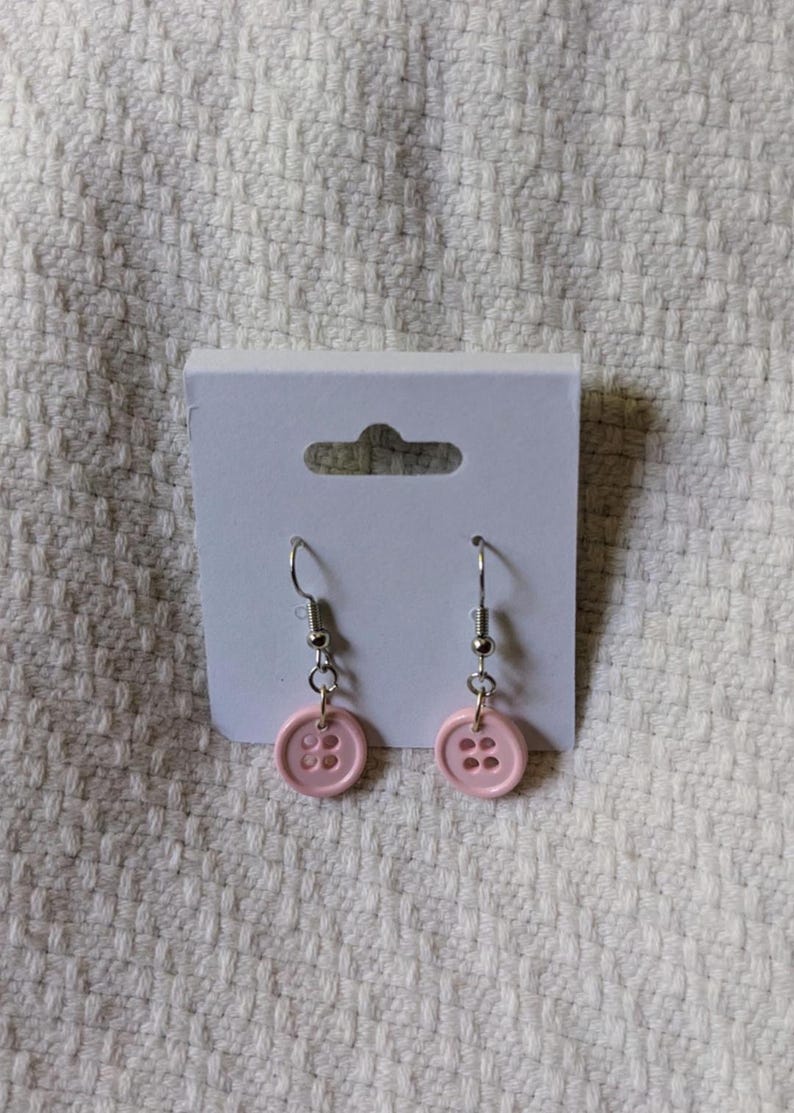 May include: A pair of button earrings. Each earring features a light pink button with four holes, suspended from a silver-colored fish hook. The earrings are displayed on a white card against a textured white background.