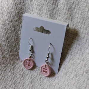 May include: A pair of pink button earrings with silver-colored hooks and chain attachments. The earrings are displayed on a white card with a small cutout at the top, set against a textured white background.